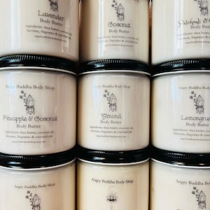 May include: Nine glass jars of body butter with labels for different scents, including Lavender, Coconut, Patchouli, Pineapple & Coconut, Almond, Lemongrass, Sandalwood & Vanilla, Dragon's Blood, and Blueberry. Each jar has a crystal design and the text "Angry Buddha Body Shop".