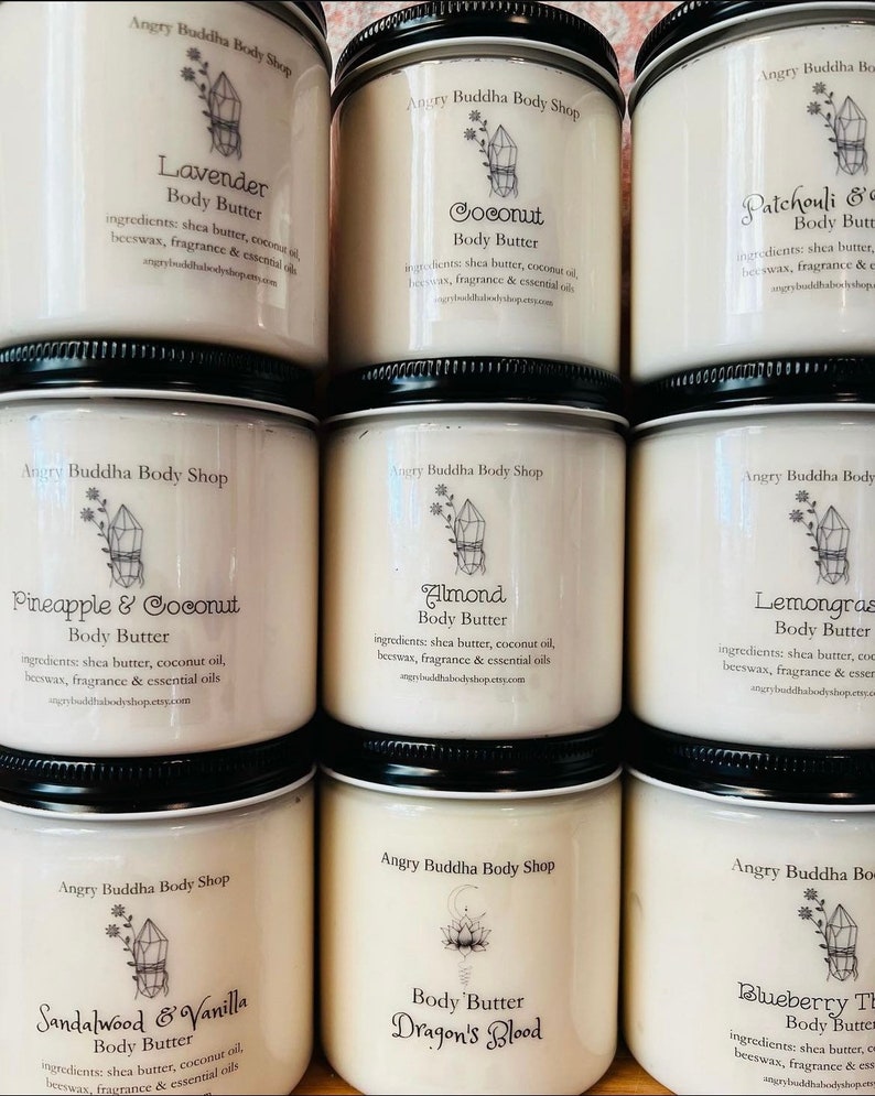 May include: Nine glass jars of body butter with labels for different scents, including Lavender, Coconut, Patchouli, Pineapple & Coconut, Almond, Lemongrass, Sandalwood & Vanilla, Dragon's Blood, and Blueberry. Each jar has a crystal design and the text "Angry Buddha Body Shop".