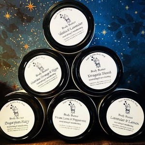 May include: Five white jars with black lids and labels. Each jar contains body butter with a different scent. The scents are: Vanilla & Lavender, Jasmine, Orange & Rose, Sugarplum Fairy, Dragon's Blood, Peace, Love & Peppermint, and Lavender & Lemon. The labels are decorated with a crystal graphic and the text "Angry Buddha Body Shop".