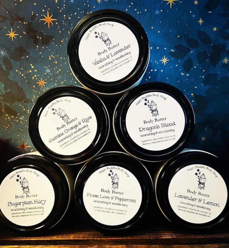 May include: Five white jars with black lids and labels. Each jar contains body butter with a different scent. The scents are: Vanilla & Lavender, Jasmine, Orange & Rose, Sugarplum Fairy, Dragon's Blood, Peace, Love & Peppermint, and Lavender & Lemon. The labels are decorated with a crystal graphic and the text "Angry Buddha Body Shop".