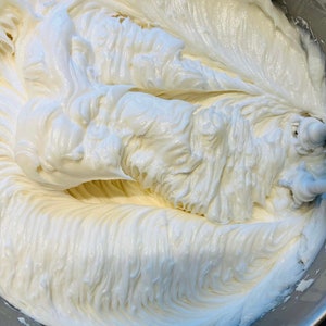 May include: A close-up of a white frosting being whipped in a silver mixing bowl. The frosting is smooth and creamy, with a slightly glossy finish.