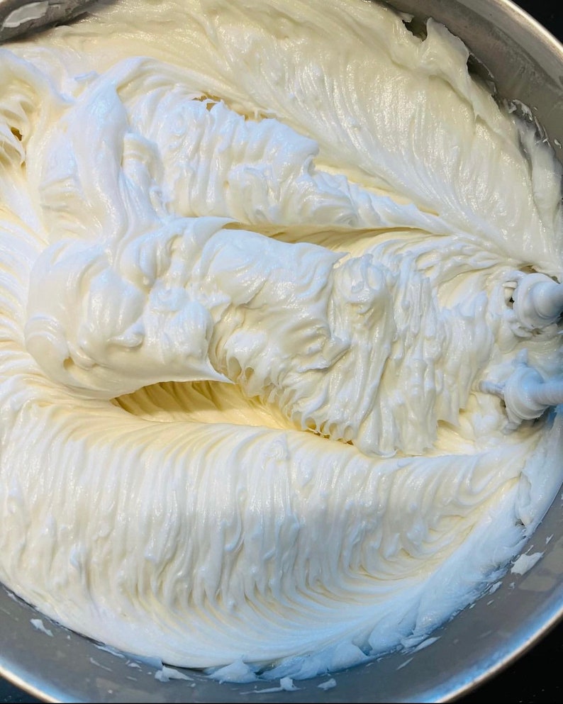 May include: A close-up of a white frosting being whipped in a silver mixing bowl. The frosting is smooth and creamy, with a slightly glossy finish.