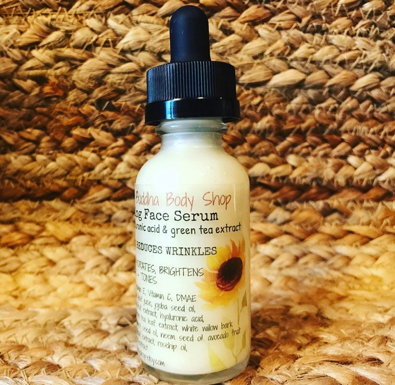 May include: A clear glass bottle with a white label that reads "Budha Body Shop Aging Face Serum Hyaluronic acid & green tea extract Reduces wrinkles Hydrates, brightens skin tones Vitamin E, Vitamin C, DMAE Aloe vera, jojoba seed oil, tea leaf extract, hyaluronic acid, white willow bark, neem seed oil, avocado fruit extract, rosehip oil."