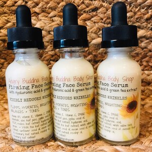 May include: Three glass bottles of a white face serum with black dropper tops. The bottles are labeled "Angry Buddha Body Shop Firming Face Serum with Hyaluronic acid & green tea extract" and list ingredients. The bottles are sitting on a woven brown surface.