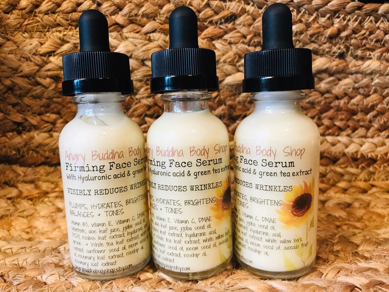 May include: Three glass bottles of a white face serum with black dropper tops. The bottles are labeled "Angry Buddha Body Shop Firming Face Serum with Hyaluronic acid & green tea extract" and list ingredients. The bottles are sitting on a woven brown surface.