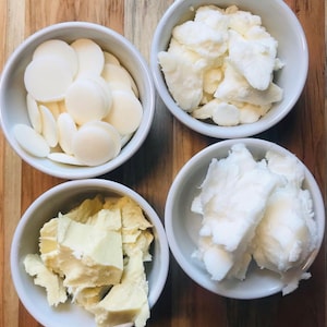 May include: Four white bowls, each containing a different type of white, solid, unrefined fat. The bowls are arranged in a square on a wooden surface.