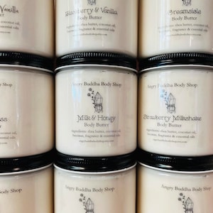 May include: A collection of eight white jars with black lids, each containing a different scented body butter. The jars are labeled with the names of the scents, including "Vanilla", "Blackberry & Vanilla", "Creamsicle", "Milk & Honey", "Strawberry Milkshake", "Flower Child", and "Island Gardenia". Each jar also includes the text "Angry Buddha Body Shop" and a small crystal graphic.
