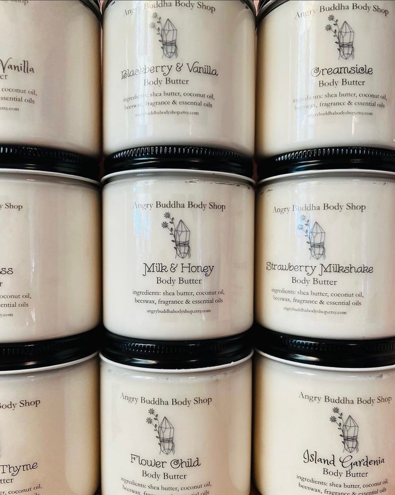 May include: A collection of eight white jars with black lids, each containing a different scented body butter. The jars are labeled with the names of the scents, including "Vanilla", "Blackberry & Vanilla", "Creamsicle", "Milk & Honey", "Strawberry Milkshake", "Flower Child", and "Island Gardenia". Each jar also includes the text "Angry Buddha Body Shop" and a small crystal graphic.