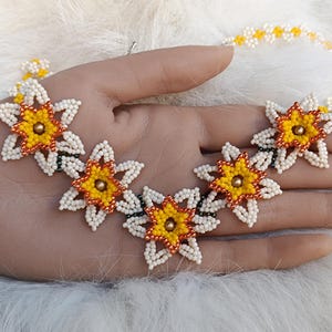 Puede incluir: Un collar de cuentas con un diseño floral. El collar tiene cuentas blancas, amarillas, naranjas y doradas. Las flores son el punto focal, con un diseño detallado de pétalos y una cuenta dorada central. El collar se muestra en una mano.