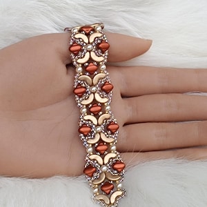 Könnte beinhalten: Ein Armband mit Perlen, das ein sich wiederholendes Muster aus rostfarbenen und goldfarbenen Perlen aufweist. Das Armband ist mit kleinen, perlenartigen Perlen akzentuiert. Der Verschluss ist silberfarben. Das Armband wird auf einer weißen, flauschigen Oberfläche präsentiert.
