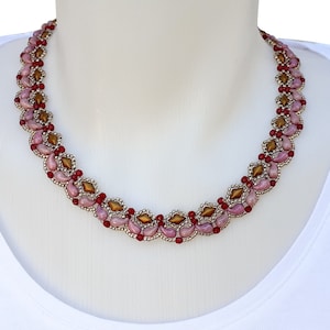 Può includere: Una collana di perline con un motivo ripetitivo di perline rosa a forma di goccia, perline color bronzo a forma di diamante e piccole perline sferiche rosse, tutte collegate tra loro con minuscole perline metalliche argentate in un design intrecciato.