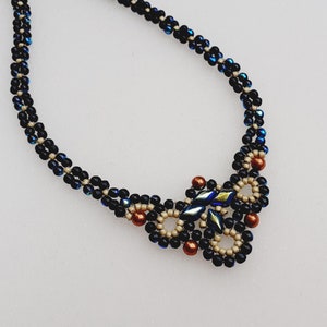 May include: A beaded necklace featuring a double-strand chain of small, iridescent dark blue and black beads. The central focal point consists of a complex woven pattern of matte beige, metallic copper, and elongated, shimmering blue-green beads.