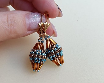 Könnte beinhalten: Ein Paar Hängeohrringe mit einem geometrischen, perlenverwebten Design. Die Ohrringe bestehen aus metallisch bronzefarbenen Stiftperlen, kleinen türkisblauen Saatperlen und goldfarbenen Brisur-Ohrhaken. Der Text Adiva Jewels Tutorials ist unten rechts zu sehen.