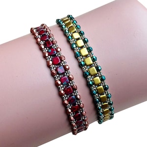 May include: Two beaded bracelets. One is red and copper-colored, featuring square and round beads. The other is gold and teal, with square gold beads and teal accents. Both are on a light pink surface.
