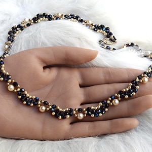 Puede incluir: Un collar con un patrón trenzado de cuentas facetadas azul oscuro y pequeñas cuentas doradas, acentuado con perlas redondas de color crema. La pieza de joyería tiene un diseño flexible y entrelazado con un cierre metálico en el extremo.