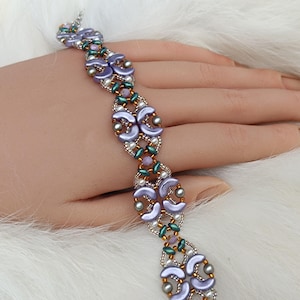 Puede incluir: Una pulsera de cuentas con cuentas color lavanda en forma de media luna, pequeñas cuentas de semillas verde metálico y diminutas perlas blancas. El diseño intrincado está tejido con hilo dorado, creando un patrón estructurado.