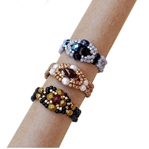 Könnte beinhalten: Drei Perlenringe mit unterschiedlich farbigen Perlen und Mittelstücken. Die Ringe sind auf einer braunen Oberfläche gestapelt.