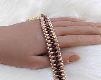 Puede incluir: Una pulsera de cuentas con un patrón repetitivo de cuentas en forma de diamante de color granate oscuro flanqueadas por cuentas doradas metálicas, bordeadas por pequeñas cuentas blancas. La pulsera incluye una cadena extensora de metal dorado.