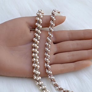 May include: Two long, flexible strands composed of small, cream-coloured spherical beads woven together with metallic rose gold accents. The strands feature a twisted, rope-like texture with a consistent pattern of alternating pearls and metallic beads.