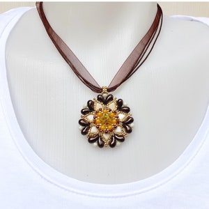 Può includere: Collana con pendente floreale e nastro marrone. Il pendente è composto da perline marroni, bianche, dorate e gialle. Il centro del pendente è composto da perline gialle e dorate.