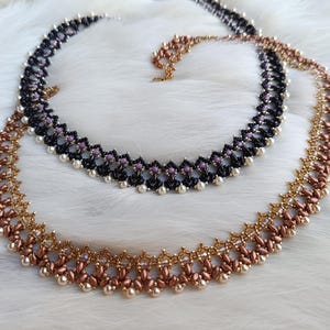 Op de afbeelding: Twee kettingen met kralen. Eén is zwart met paarse en witte accenten, de andere is koper en goud. Beide kettingen hebben een delicaat, ingewikkeld ontwerp, met kleine kralen die een herhalend patroon vormen. De kettingen worden gepresenteerd op een witte, zachte ondergrond.