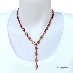 以下が含まれることがあります： Y字型のペンダントが付いた、銅色のビーズネックレス。ネックレスは、小さくて丸いビーズと、大きくて楕円形のビーズが特徴です。ペンダントは、先端に向かって細くなる一連の楕円形のビーズを持っています。ネックレスは白い背景に表示されています。