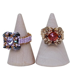 May include: Two beaded rings on wooden display stands. The ring on the left has a pink and purple beaded design with a clear crystal in the center. The ring on the right has a gold and brown beaded design with a red crystal in the center.