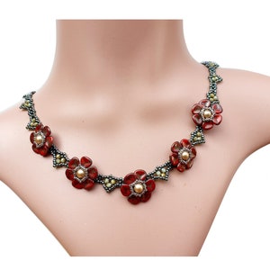 May include: A beaded necklace featuring five red flower-shaped beads with gold-toned centers. The flowers are connected by dark grey seed beads and small clusters of light green beads, creating a floral pattern along the entire length.