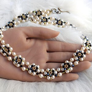Può includere: Una collana di perline con un motivo floreale. La collana presenta file alternate di perle bianche e perline a forma di fiore nere e dorate. La chiusura è dorata. La collana è esposta su una superficie bianca e soffice.