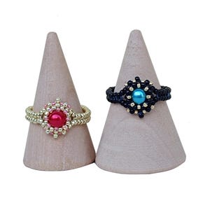 Könnte beinhalten: Zwei auf Holzkonen präsentierte Perlenringe. Ein Ring hat ein goldenes Band mit einem roten Mittelstein, der andere ein schwarzes Band mit einem blauen Mittelstein. Die Ringe sind handgefertigt und zeigen aufwendige Perlenarbeiten.