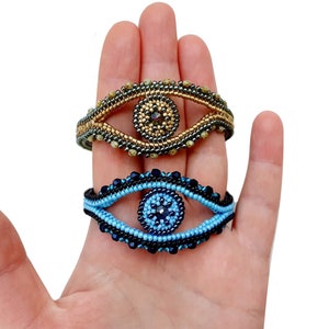 Puede incluir: Dos pulseras de cuentas con un diseño de ojo turco. La pulsera superior es dorada y negra con un centro azul oscuro. La pulsera inferior es azul y negra con un centro azul claro.