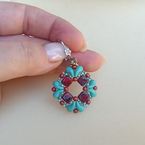 May include: A handmade earring featuring a turquoise and red beaded design. The earring has a circular shape with a square center and is held by a silver hook. The beads are small and intricately arranged, creating a detailed and colorful accessory.