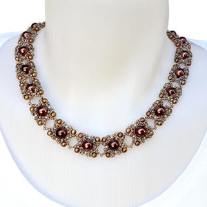 Peut inclure: Un collier de perles composé d'un motif répétitif de perles sphériques marron foncé entourées de petites perles bronze métallisées et de grappes de perles de rocaille tissées, formant un design structuré et symétrique autour du cou.