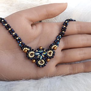 May include: A beaded necklace in dark blue and gold. The necklace has a floral pendant and is displayed on a hand. The beads are small and the design is intricate.