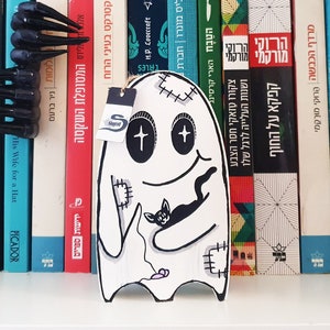 Puede incluir: Un fantasma blanco con ojos negros y una boca cosida sostiene un gato negro. El fantasma est&aacute; en una estanter&iacute;a con una variedad de libros en diferentes idiomas.