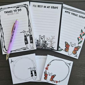 May include: A collection of spooky-themed stationery items. Includes lined notepads with phrases like "Things to Do Before I Die" and "I'll Rest in My Grave." Also features square sticky notes and a purple pen. The designs include ghost illustrations and a butterfly.
