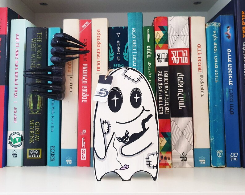 Puede incluir: Un fantasma blanco con ojos negros y una boca cosida sostiene un gato negro. El fantasma est&aacute; en una estanter&iacute;a con una variedad de libros en diferentes idiomas.