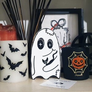 Puede incluir: Un fantasma blanco con costuras cosidas y un gato negro en sus brazos. El fantasma tiene grandes ojos negros y una sonrisa cosida. El fantasma est&aacute; de pie sobre una superficie negra con una vela negra y blanca, una vela negra con un dise&ntilde;o de telara&ntilde;a y calabaza, y un candelabro negro con un dise&ntilde;o circular.