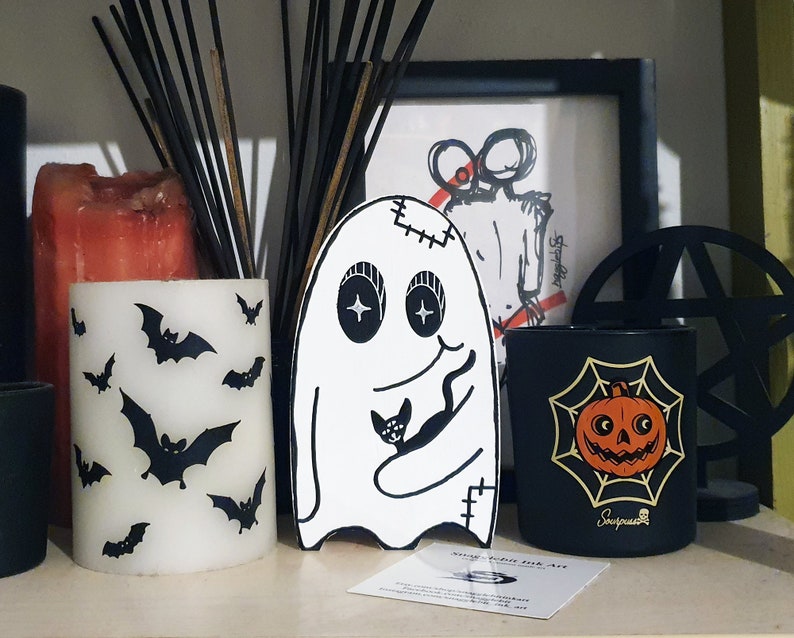 Puede incluir: Un fantasma blanco con costuras cosidas y un gato negro en sus brazos. El fantasma tiene grandes ojos negros y una sonrisa cosida. El fantasma est&aacute; de pie sobre una superficie negra con una vela negra y blanca, una vela negra con un dise&ntilde;o de telara&ntilde;a y calabaza, y un candelabro negro con un dise&ntilde;o circular.