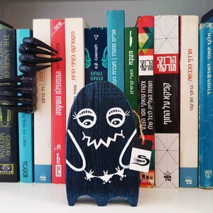 Puede incluir: Una figurita de fantasma de madera azul con ojos, boca y una peque&ntilde;a criatura pintados de blanco en la parte inferior. El fantasma est&aacute; de pie en una estanter&iacute;a con una variedad de libros en hebreo e ingl&eacute;s.