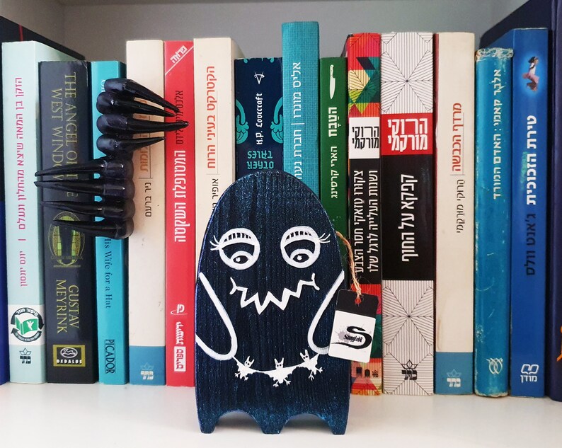 Puede incluir: Una figurita de fantasma de madera azul con ojos, boca y una peque&ntilde;a criatura pintados de blanco en la parte inferior. El fantasma est&aacute; de pie en una estanter&iacute;a con una variedad de libros en hebreo e ingl&eacute;s.