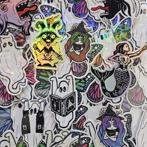 Pode incluir: Uma coleção de autocolantes coloridos com designs assustadores e caprichosos. Os autocolantes incluem fantasmas, uma sereia, uma criatura semelhante a uma cabra e uma bruxa, todos com um acabamento holográfico. Padrões de asas de borboleta também são visíveis.