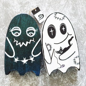 Puede incluir: Dos adornos de fantasmas de madera, uno verde con murci&eacute;lagos y el otro blanco con un gato negro. Ambos fantasmas tienen detalles cosidos y ojos negros.