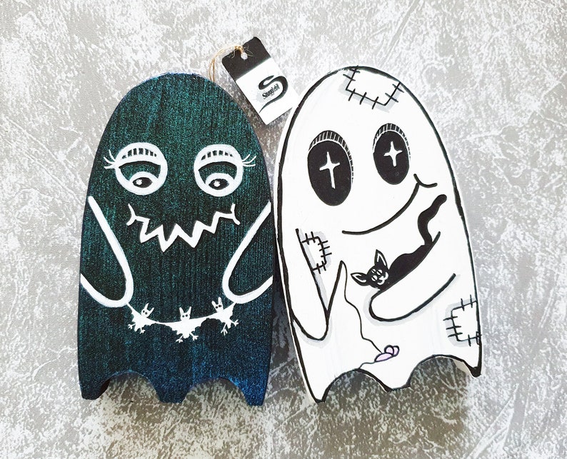Puede incluir: Dos adornos de fantasmas de madera, uno verde con murci&eacute;lagos y el otro blanco con un gato negro. Ambos fantasmas tienen detalles cosidos y ojos negros.