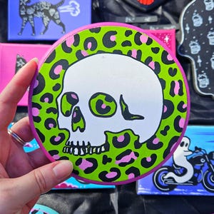 Może przedstawiać: Okrągłe, ręcznie malowane płótno przedstawiające białą czaszkę z zielonymi i różowymi akcentami. Czaszka otoczona jest zieloną obwódką w panterkę z różowymi plamkami, osadzoną na różowej krawędzi. Inne małe płótna są w tle.