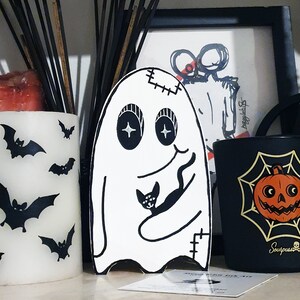 Puede incluir: Tres velas negras con un fantasma blanco, una vela negra con una telara&ntilde;a y una calabaza, y una vela blanca con murci&eacute;lagos. Las velas est&aacute;n sobre una superficie de madera.