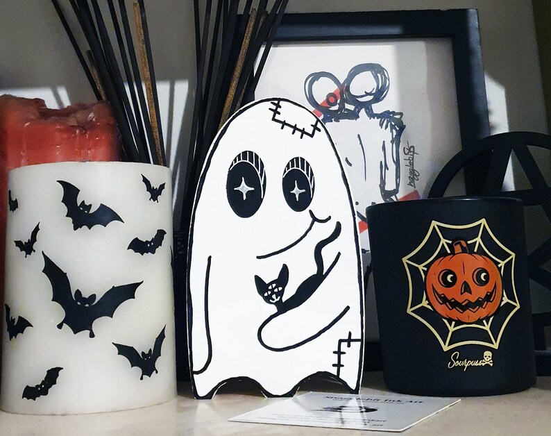 Puede incluir: Tres velas negras con un fantasma blanco, una vela negra con una telara&ntilde;a y una calabaza, y una vela blanca con murci&eacute;lagos. Las velas est&aacute;n sobre una superficie de madera.