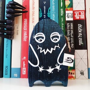 Puede incluir: Una figurita de fantasma de madera azul con ojos, boca y murci&eacute;lagos pintados de blanco colgando de su cuello. El fantasma est&aacute; de pie en una estanter&iacute;a con una etiqueta que dice "Stangibl".