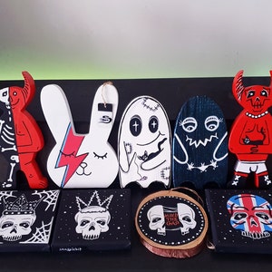 Puede incluir: Una colecci&oacute;n de ocho figuritas de madera pintadas a mano y obras de arte de pared. Las figuritas incluyen un diablo rojo, un conejito blanco, un fantasma y un monstruo azul. Las obras de arte de pared presentan calaveras con coronas y una calavera con el texto "Mind the Gap".