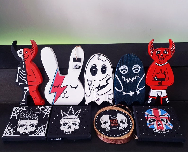 Puede incluir: Una colecci&oacute;n de ocho figuritas de madera pintadas a mano y obras de arte de pared. Las figuritas incluyen un diablo rojo, un conejito blanco, un fantasma y un monstruo azul. Las obras de arte de pared presentan calaveras con coronas y una calavera con el texto "Mind the Gap".